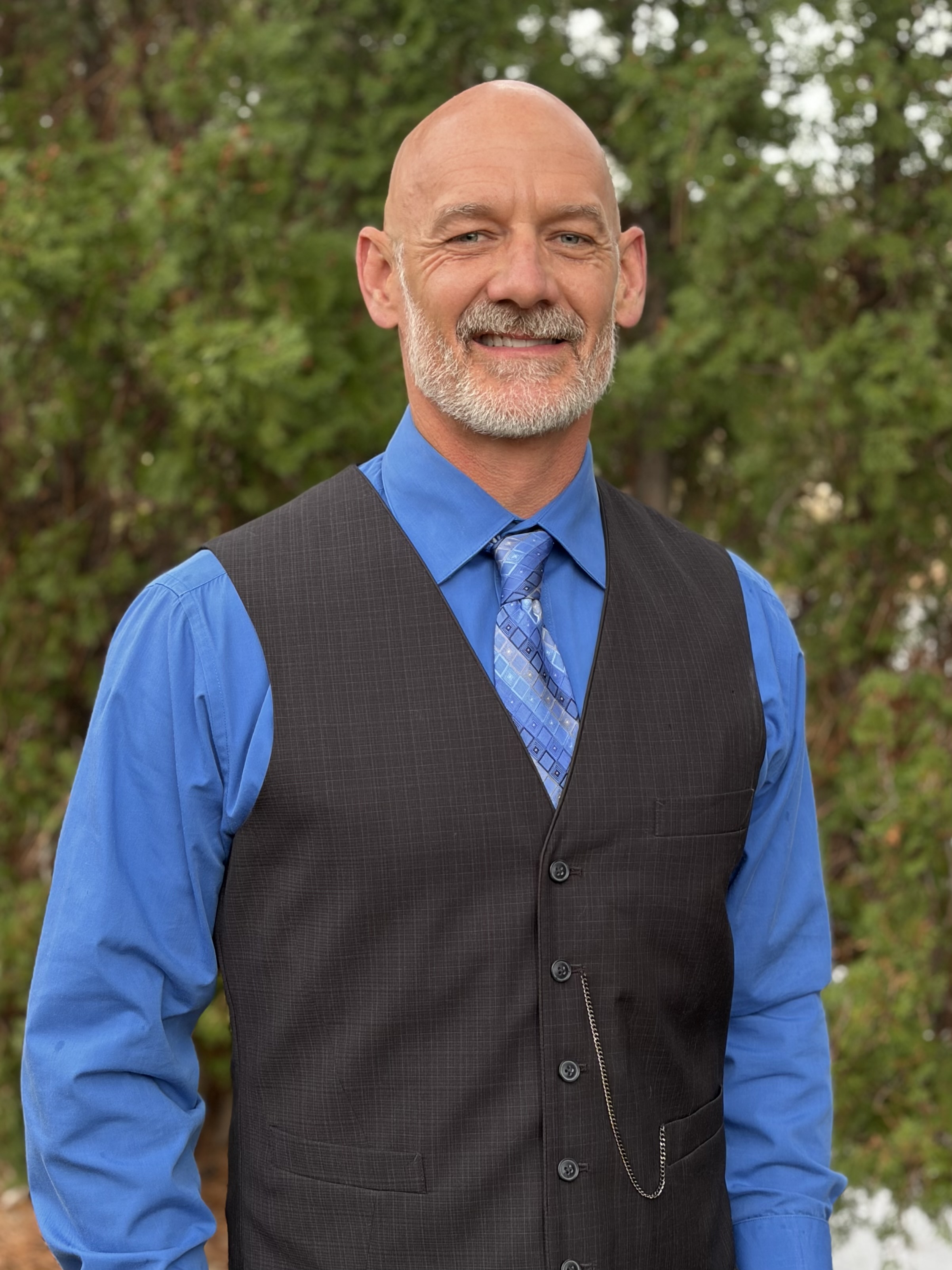 A smiling man dressed in a blue shirt and a dark vest with a tie, standing outdoors against a backdrop of greenery.